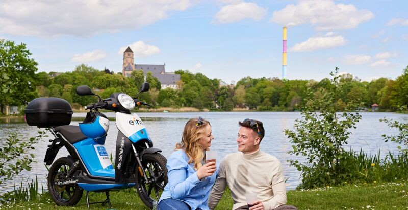 junges Paar sitzt vor Schlossteich, E-Roller steht daneben, Esse Chemnitz im Hintergrund