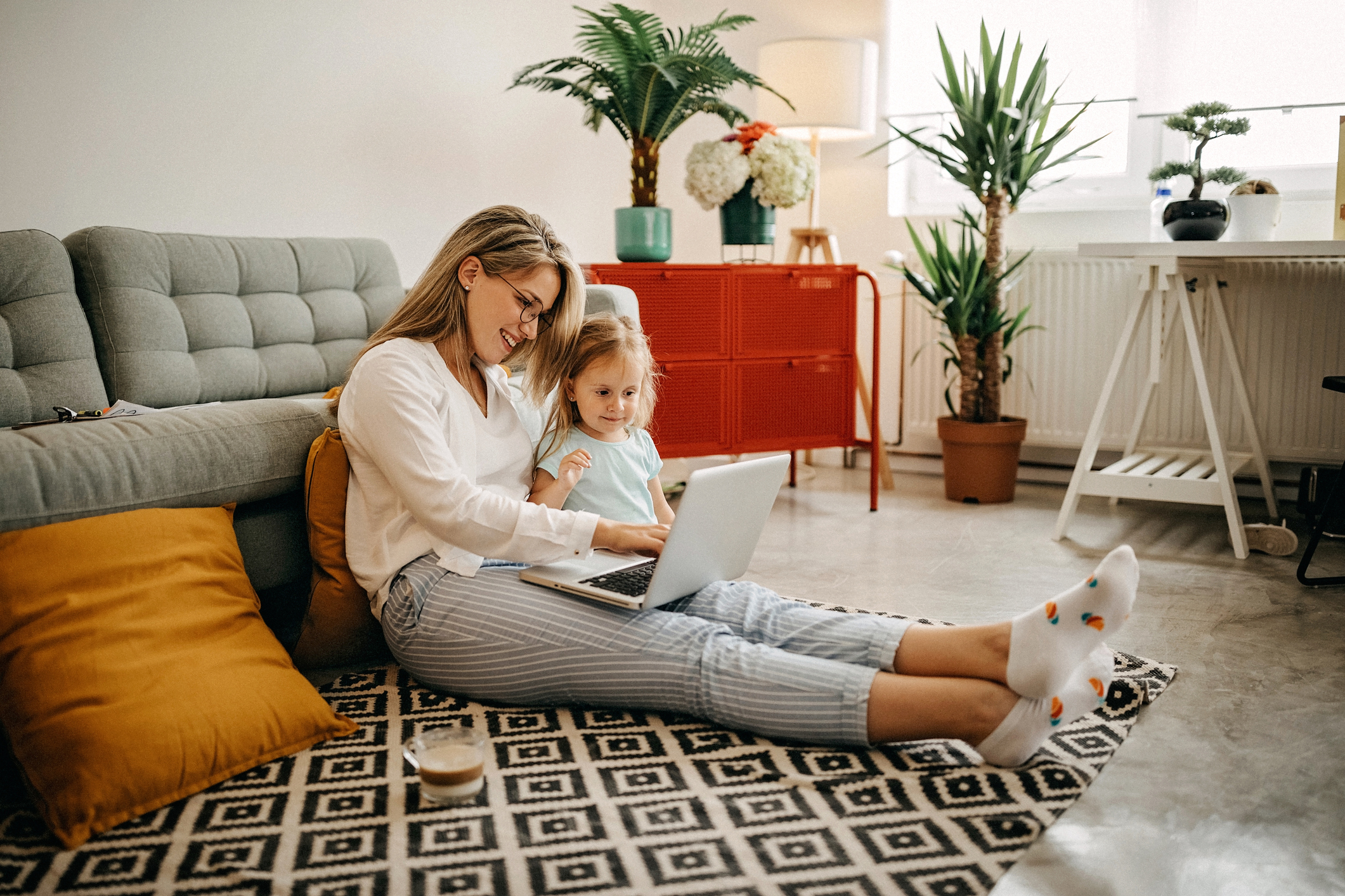 Mutter und kleine Tochter sitzen auf dem Wohnzimmer-Teppich und schauen gemeinsam in den Laptop