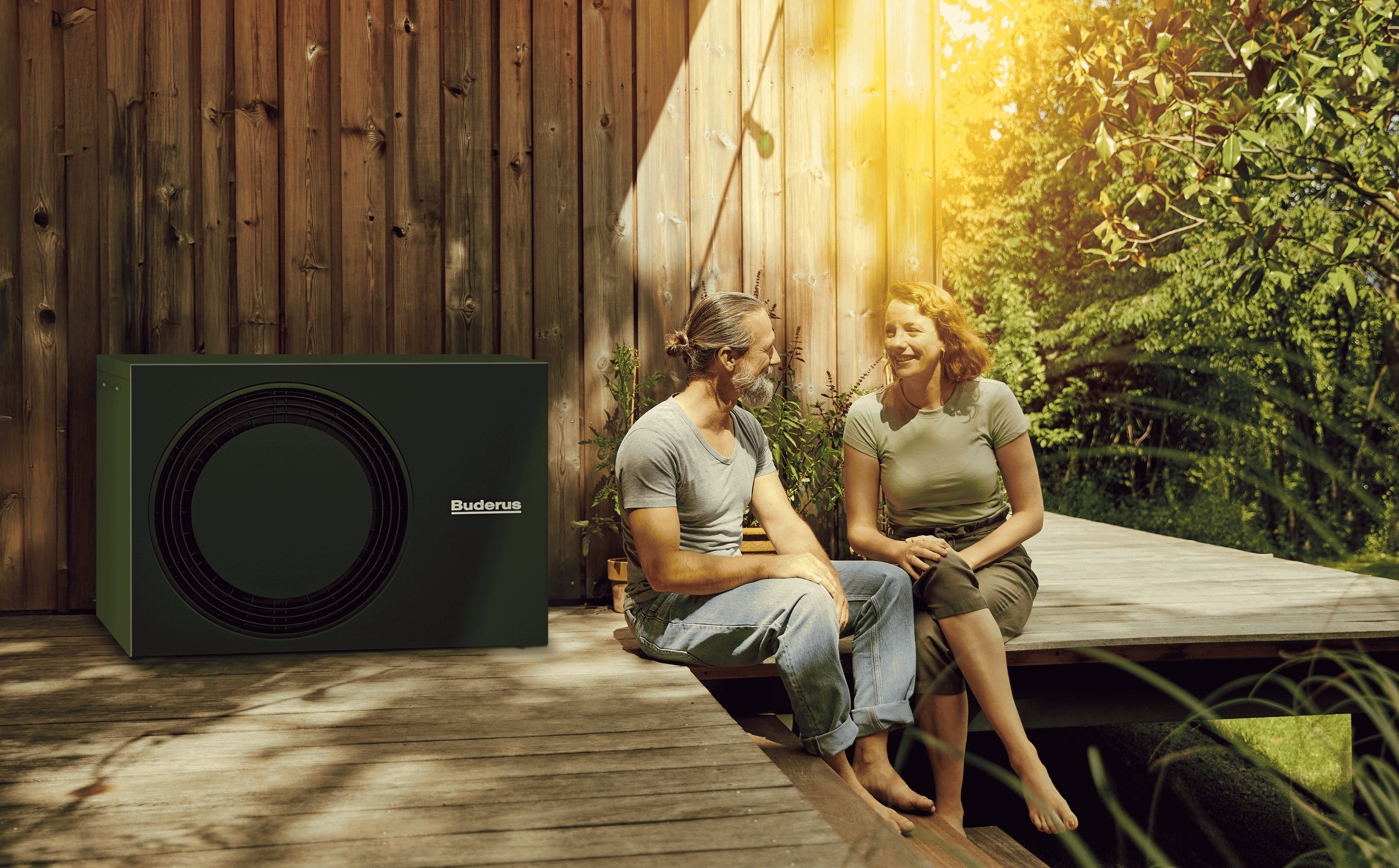 Ein Mann und eine Frau sitzen auf ihrer Holzterrasse vor ihrem Haus, neben ihnen steht eine Wärmepumpe von Bruderus