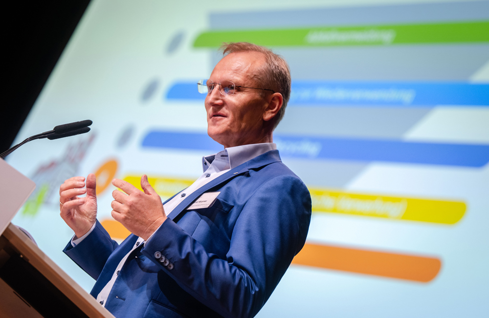 Roland Warner steht am Rednerpult und hält einen Vortag. Hinter ihm sieht man eine PowerPoint Präsentation.