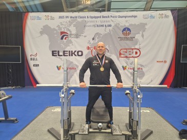 Lars Bretfeld steht stolz hinter seiner Hantelbank und blickt in die Kamera. Er trägt seine Bronzemedaille um den Hals.