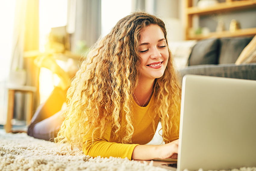 Junge Frau liegt mit dem Laptop auf dem Teppich und strahlt zufrieden.