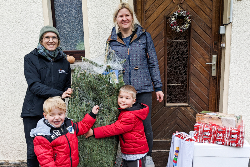 Eine Mitarbeiterin der eins und eine Familie. Die eins-Mitarbeiterin übergibt der Familie einen Tannenbaum.