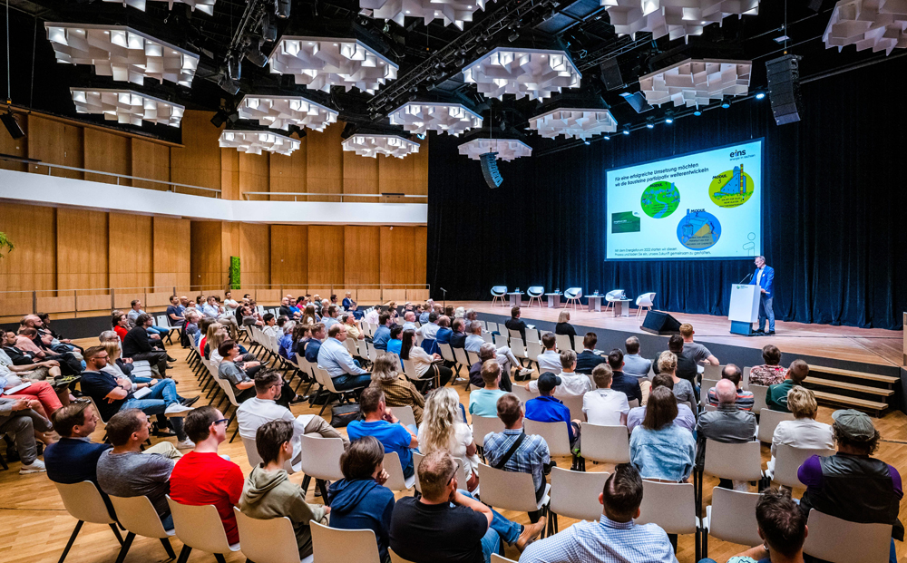 Ein Redner hält eine Rede vor einer großen Gruppe Mitarbeitern. Hinter ihm sieht man eine PowerPoint Präsentation.