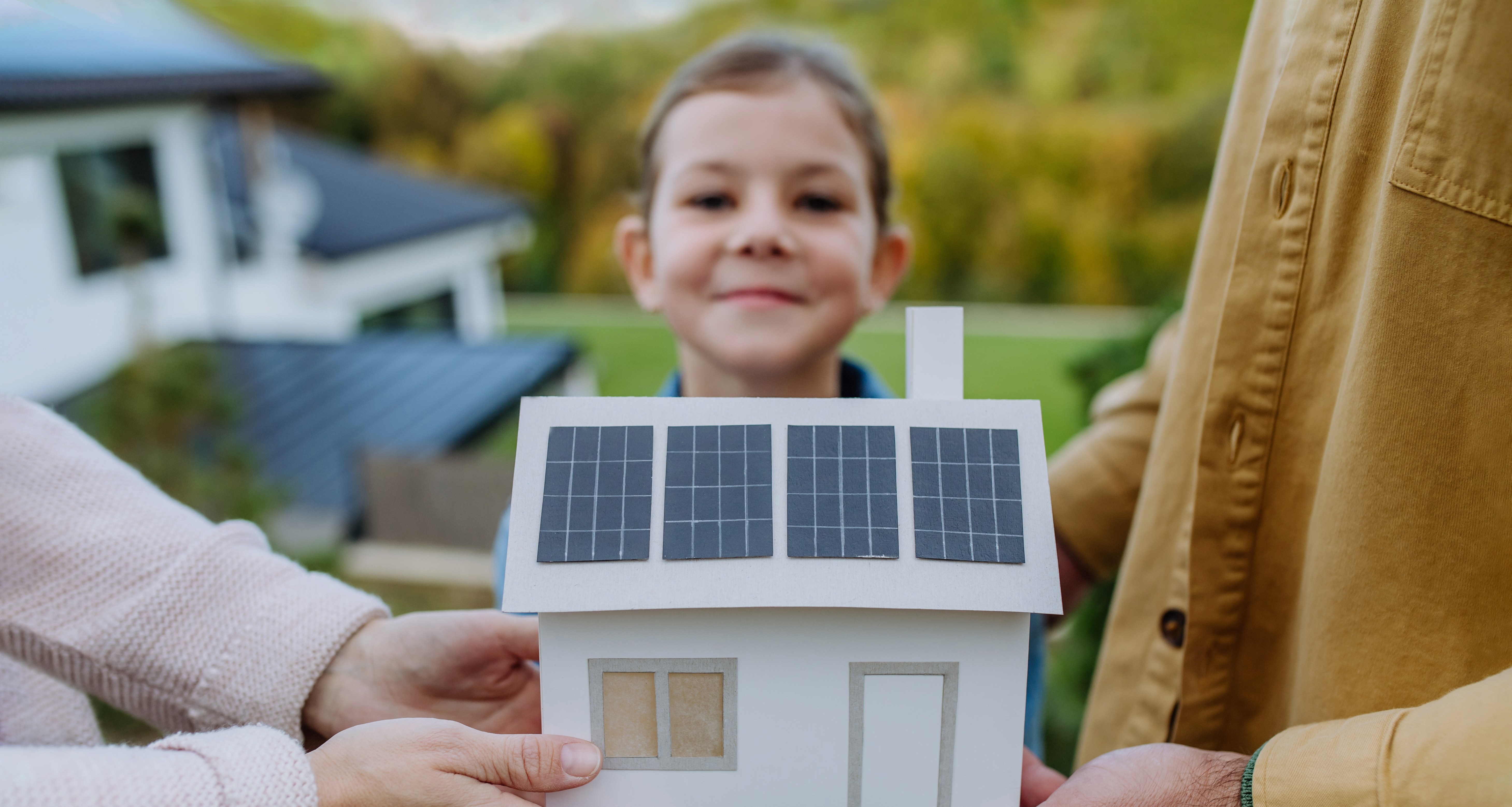 ein Mädchen mit stolzem Gesichtsausdruck hält gemeinsam mit ihren Eltern ein Papier-Hausmodell mit Solardach, welches symbolisch für ihr eigenes Einfamilienhaus steht 