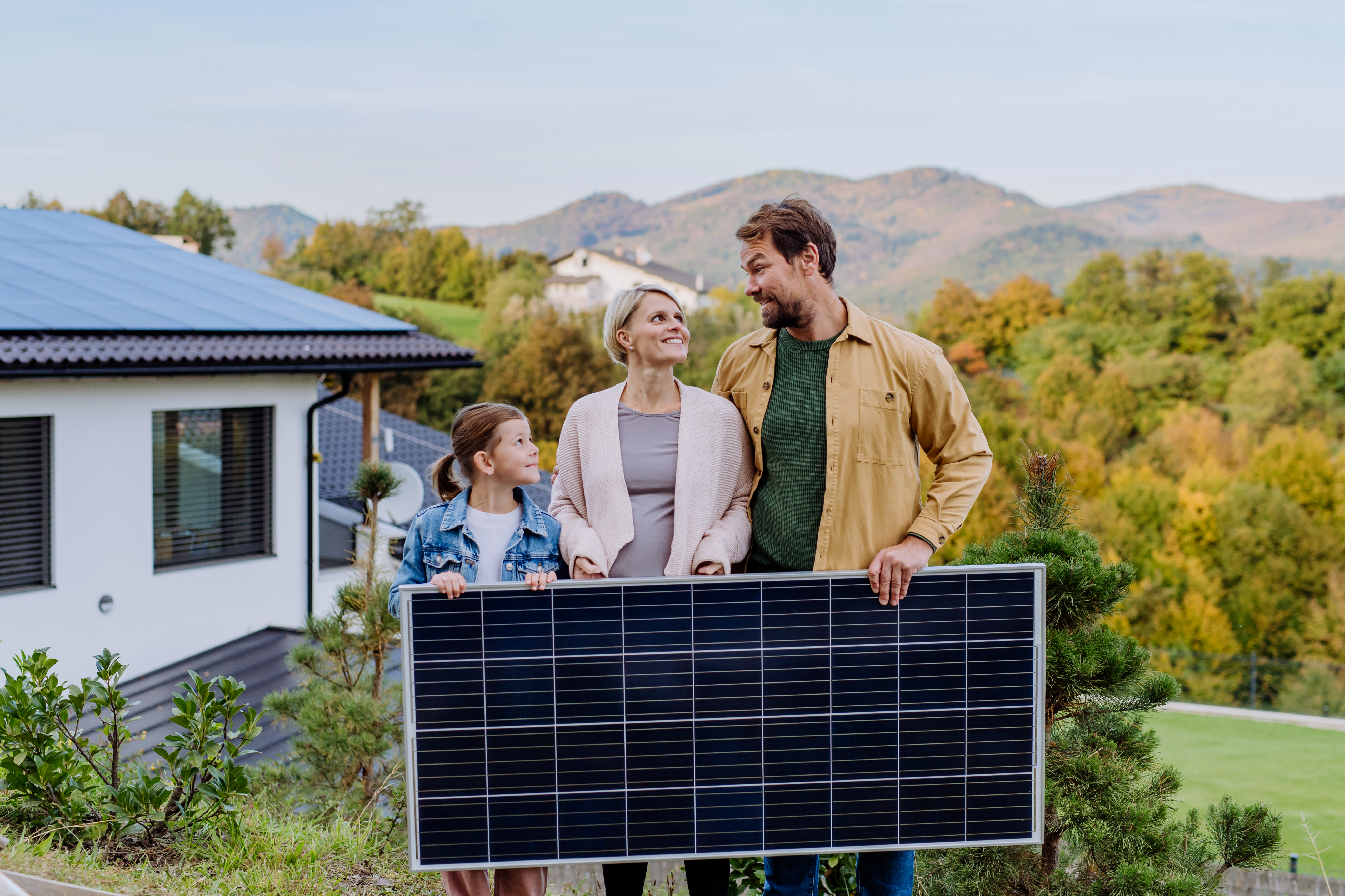 Tochter, Mutter und Vater stehen vor ihrem Einfamilienhaus und halten ein Solarpanel