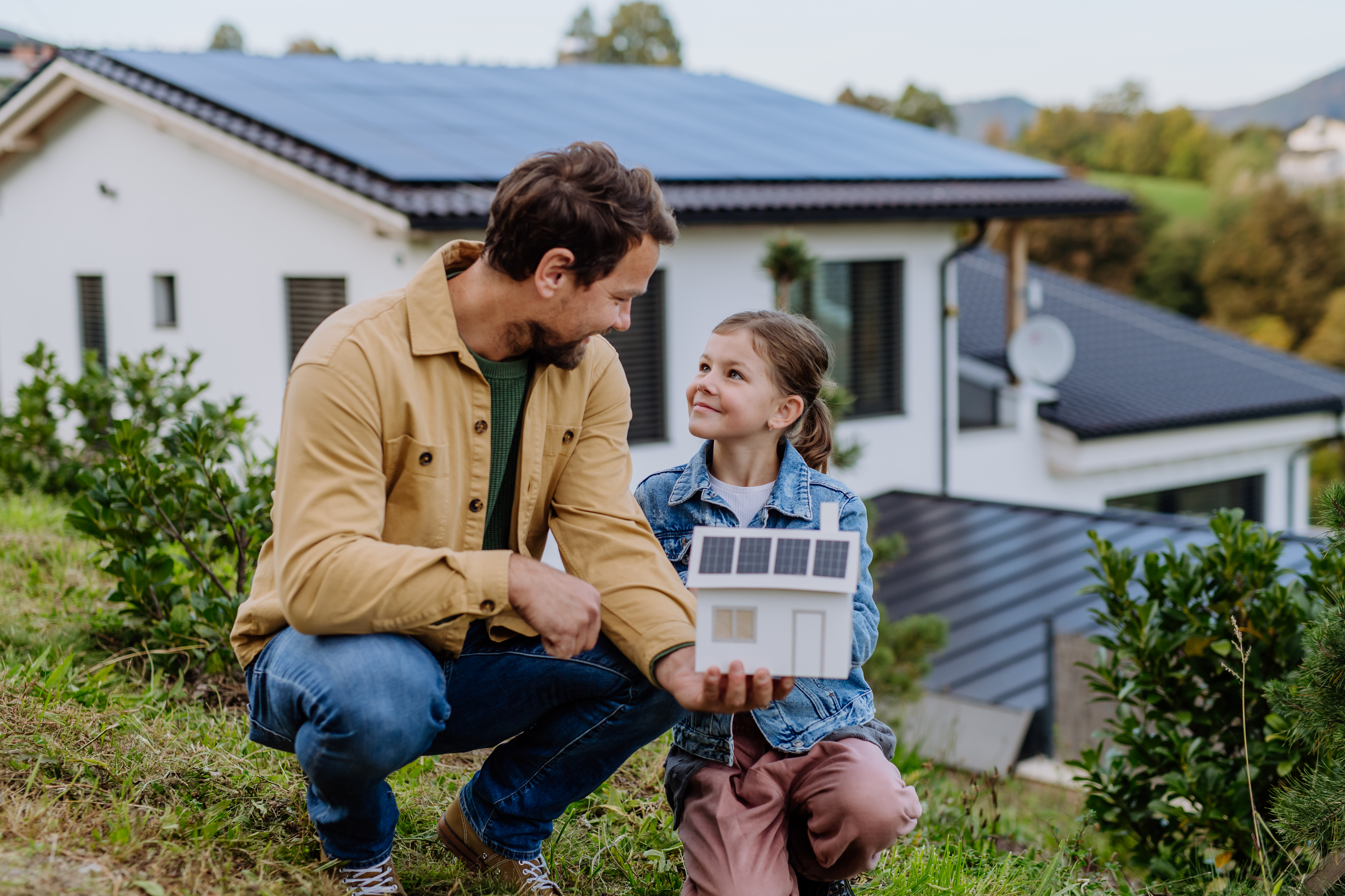 Papa und Tochter hocken vor ihrem Einfamilienhaus und betrachten ein Papier-Hausmodell mit Solardach