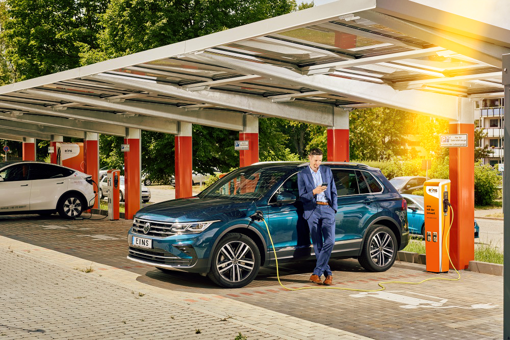 Geschäftsmann lehnt an E-Auto während dem Ladevorgang an einer Ladestation von eins