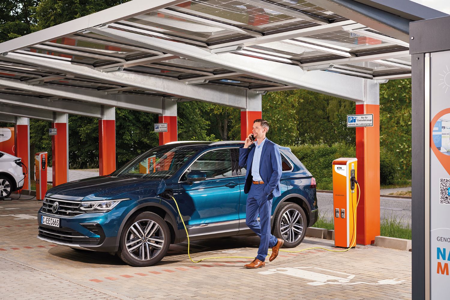 Geschäftsmann lehnt an E-Auto während dem Ladevorgang an einer Ladestation von eins