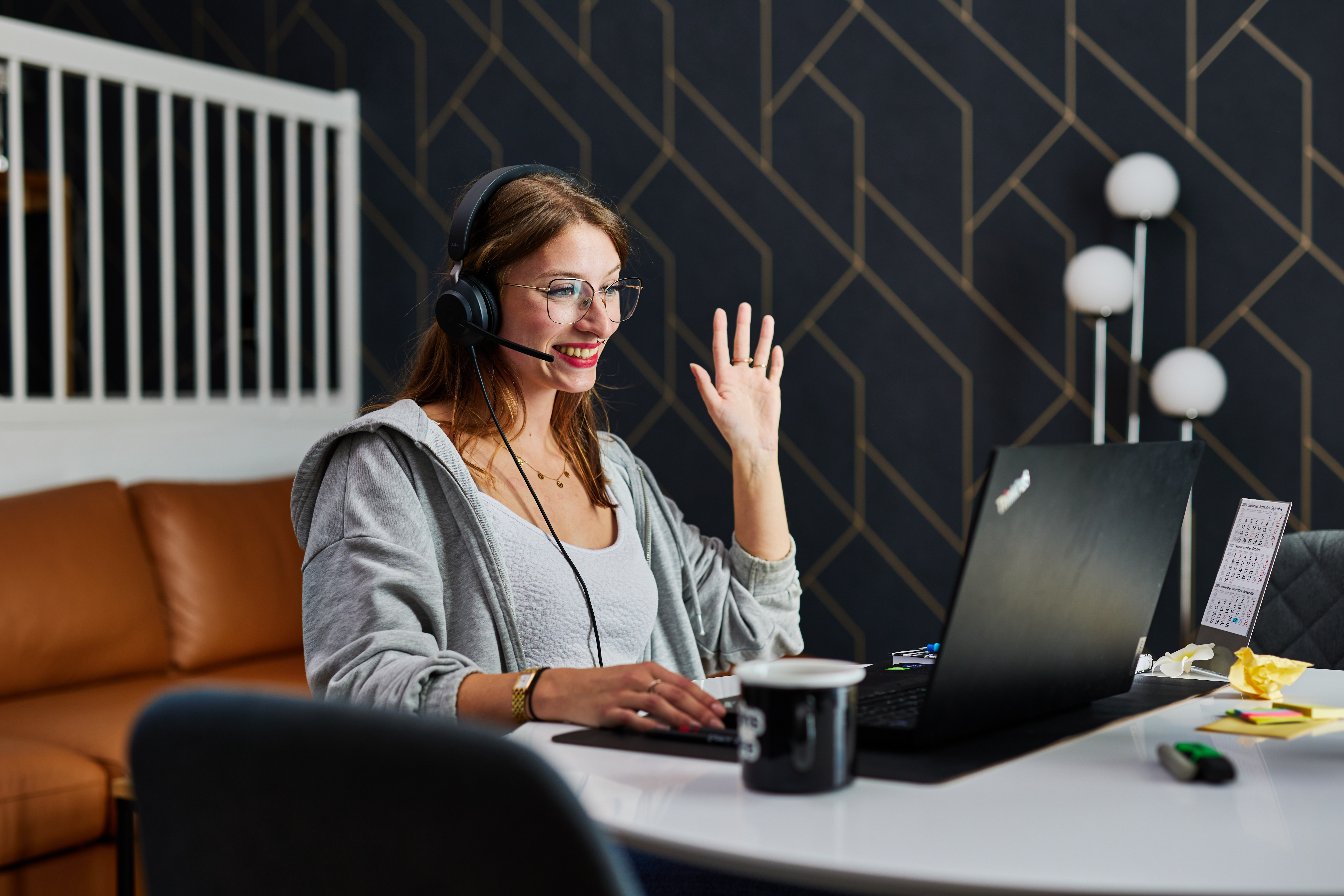 Frau mit Headsetz sitzt am Tisch und winkt in eine Laptopkamera