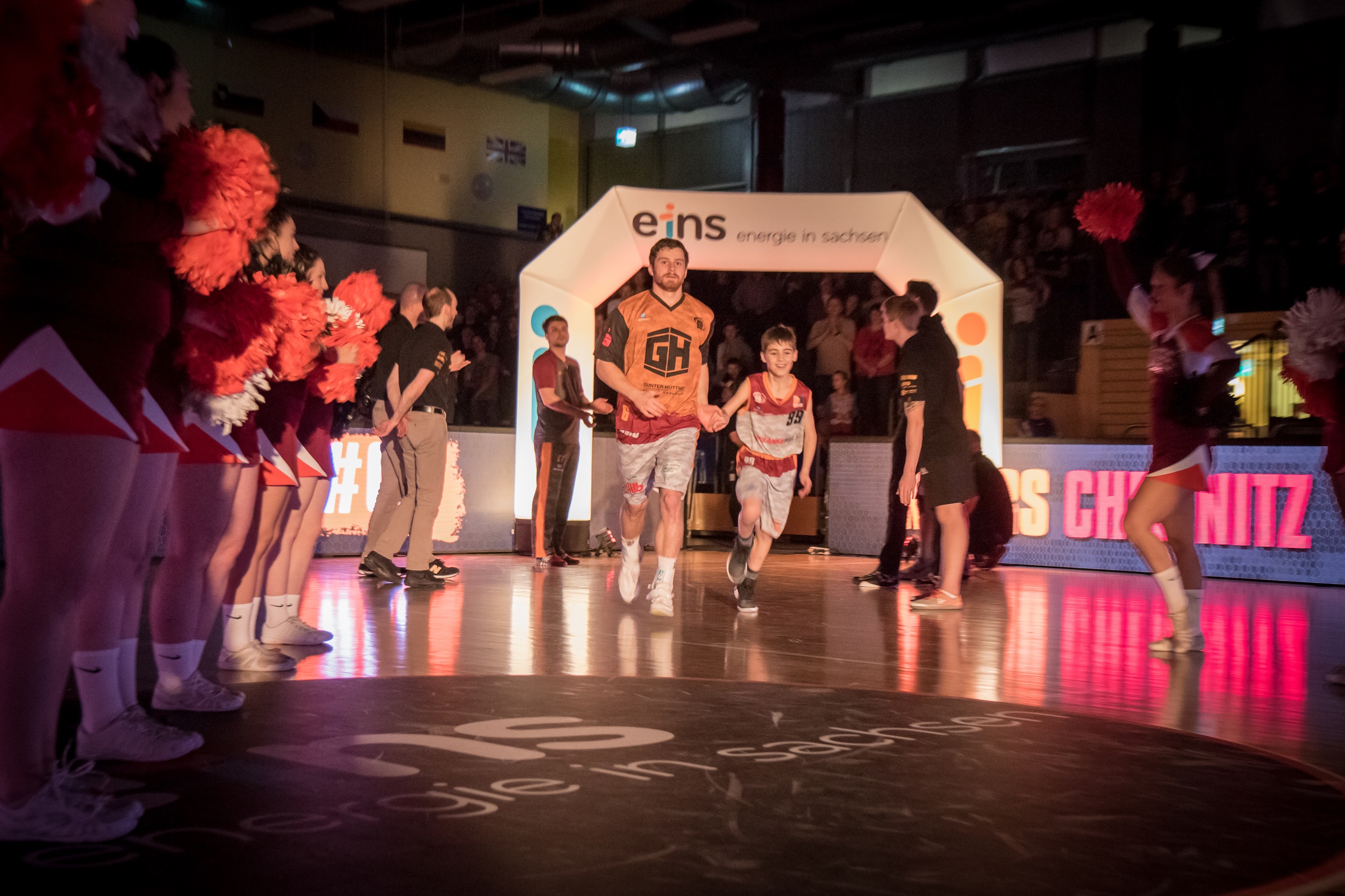 Basketbalspieler läuft ein mit Kind an der Hand umringt von Cheerleadern