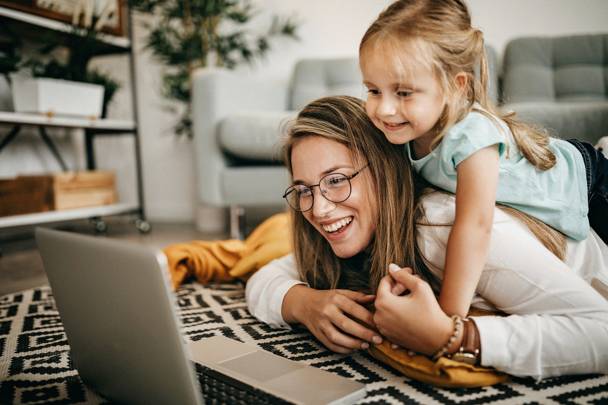 Mutter und kleine Tochter liegen auf dem Wohnzimmer-Teppich und schauen in den Laptop