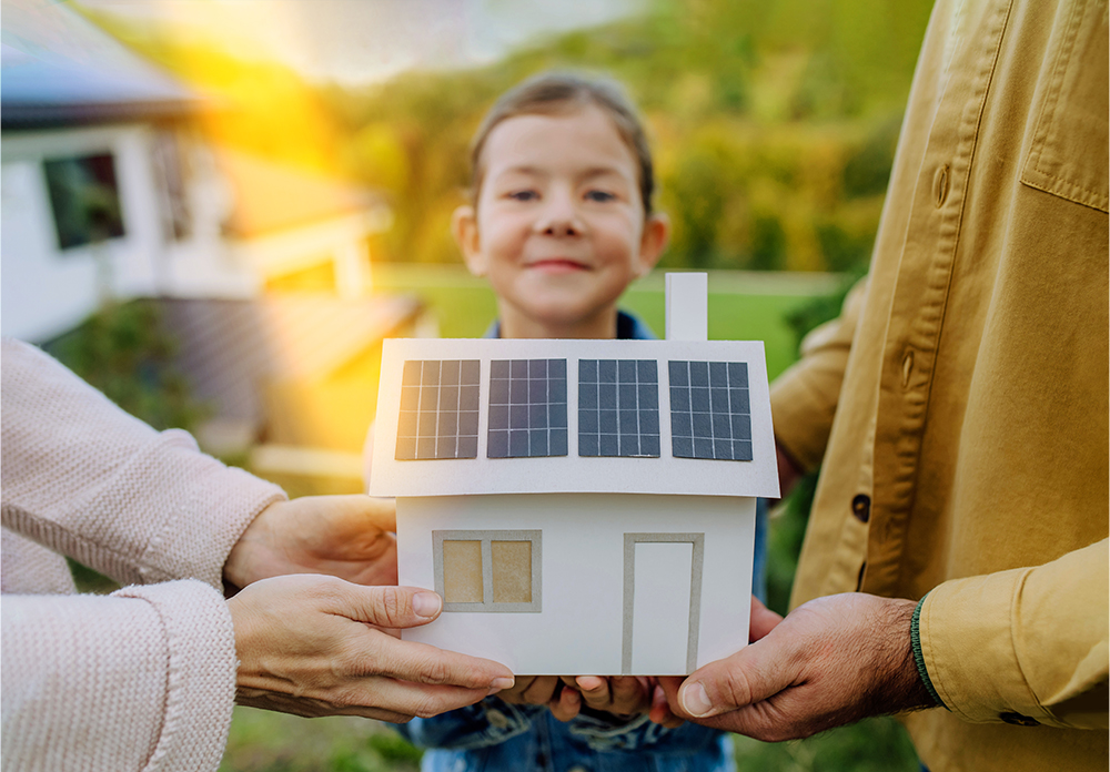 ein Mädchen mit stolzem Gesichtsausdruck hält gemeinsam mit ihren Eltern ein Papier-Hausmodell mit Solardach, welches symbolisch für ihr eigenes Einfamilienhaus steht 